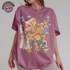 May include: A berry-colored short-sleeve t-shirt featuring a graphic print of multiple illustrated characters with blonde hair in various outfits. The design includes small blue clouds, a rocket, a yellow star, and a colorful ball. The shirt has a crew neckline.