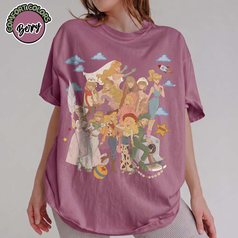 May include: A berry-colored short-sleeve t-shirt featuring a graphic print of multiple illustrated characters with blonde hair in various outfits. The design includes small blue clouds, a rocket, a yellow star, and a colorful ball. The shirt has a crew neckline.