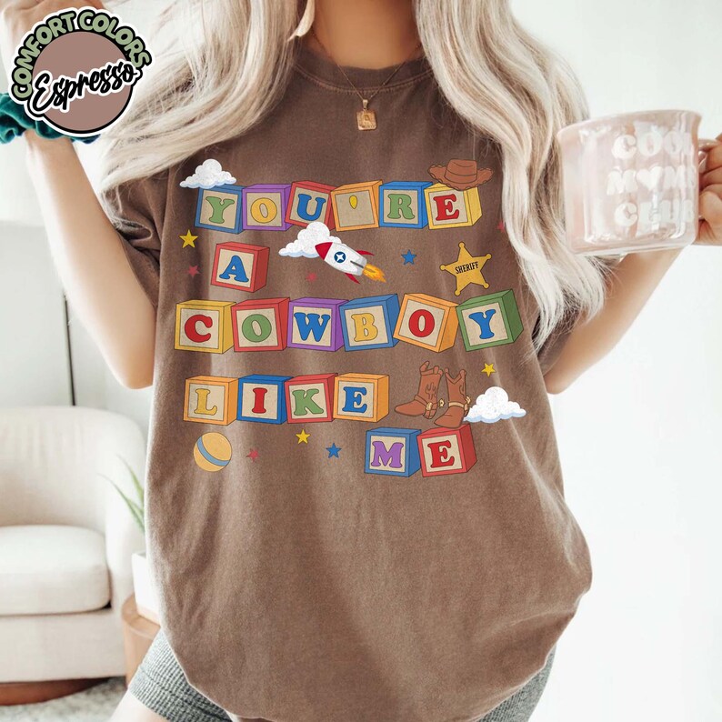 May include: A brown t-shirt featuring a graphic of colorful alphabet blocks spelling out the text YOU'RE A COWBOY LIKE ME. The design includes illustrations of a rocket ship, a sheriff star, a cowboy hat, and a pair of brown cowboy boots.