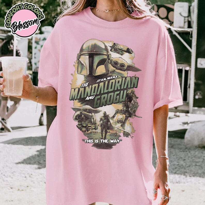 May include: A light pink t-shirt featuring a graphic print of The Mandalorian and Grogu. The design includes the text "STAR WARS THE MANDALORIAN AND GROGU" and "THIS IS THE WAY" with various character illustrations in muted green, brown, and grey tones.