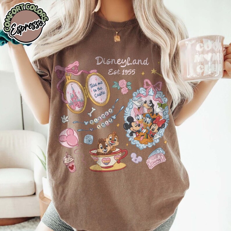 Peut inclure: T-shirt marron avec des graphismes de Disneyland. Le motif comprend un m&eacute;daillon avec un ch&acirc;teau, des personnages Disney, Tic et Tac dans une tasse &agrave; th&eacute; et le texte "Disneyland Est. 1955".
