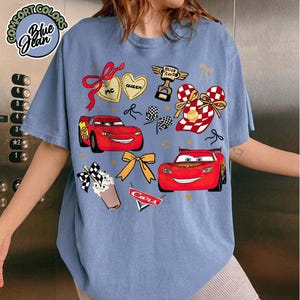 Puede incluir: Camiseta azul Comfort Colors con un dise&ntilde;o de la pel&iacute;cula Cars. El dise&ntilde;o incluye a Rayo McQueen, banderas de carreras y el texto "Cars". La camiseta tambi&eacute;n tiene el texto "Blue Jean" y "Comfort Colors".
