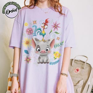May include: A light purple t-shirt featuring a cartoon pig and rooster with floral accents. The shirt has the text "COMFORT COLORS Orchid" in a circular design at the top left.