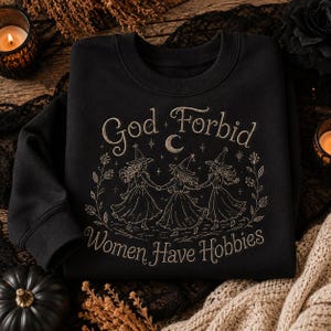 Könnte beinhalten: Ein schwarzes Sweatshirt mit Rundhalsausschnitt und einem goldfarbenen Grafikdruck von drei Figuren mit spitzen Hüten und Kleidern, die sich im Kreis an den Händen halten. Der Text lautet God Forbid Women Have Hobbies, ergänzt durch Sterne und eine Mondsichel.