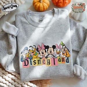 May include: A sport grey crewneck sweatshirt featuring an embroidered graphic of Disney characters including Donald Duck, Pluto, Minnie Mouse, Mickey Mouse, Daisy Duck, and Goofy above the colorful text Disney Land in a block letter font.
