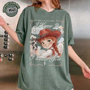 Peut inclure: Un t-shirt vert mousse avec une illustration de personnage portant un chapeau de cow-boy marron et un gant &agrave; motif vache. Le texte indique "EST 2026", "I Knew It" et "I Knew You" avec une note de musique. Le motif inclut des nuages et des &eacute;toiles.