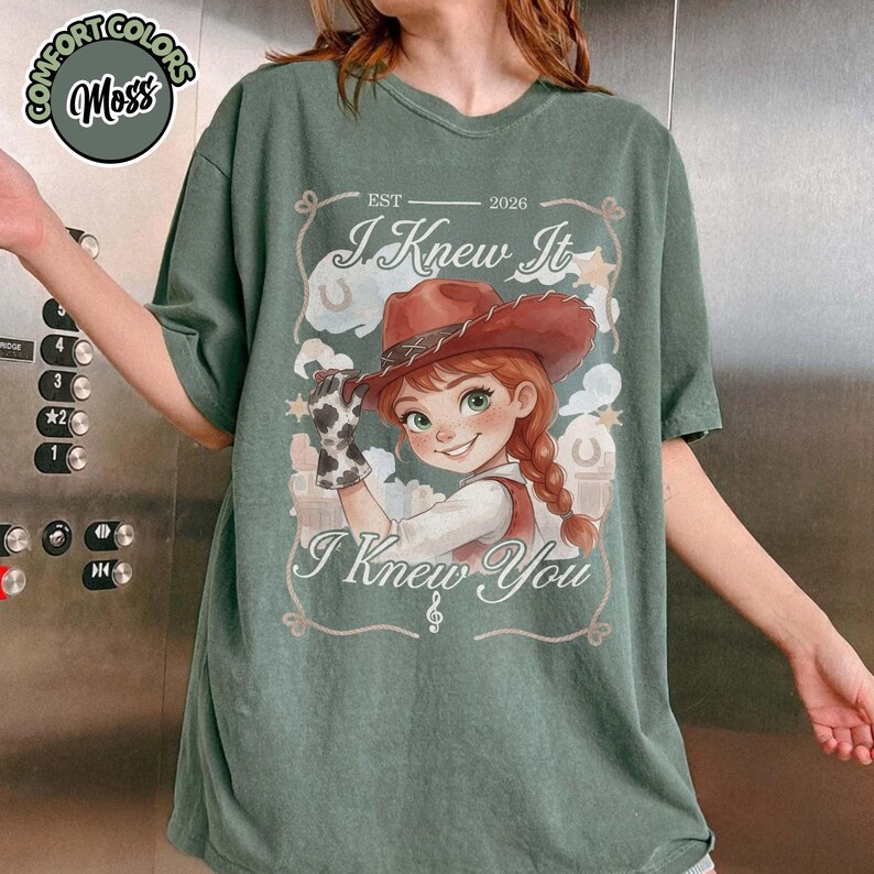 Peut inclure: Un t-shirt vert mousse avec une illustration de personnage portant un chapeau de cow-boy marron et un gant &agrave; motif vache. Le texte indique "EST 2026", "I Knew It" et "I Knew You" avec une note de musique. Le motif inclut des nuages et des &eacute;toiles.