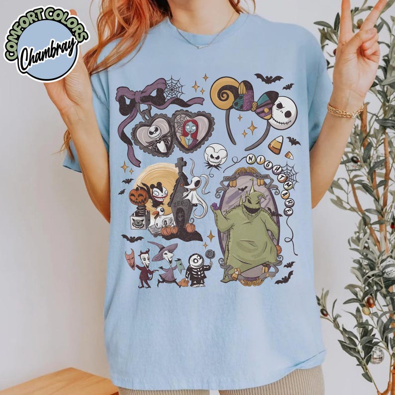 May include: Light blue t-shirt featuring a colorful graphic design with characters and elements from "The Nightmare Before Christmas." The design includes Jack Skellington, Sally, Oogie Boogie, and other iconic imagery. The shirt has a crew neck and short sleeves.