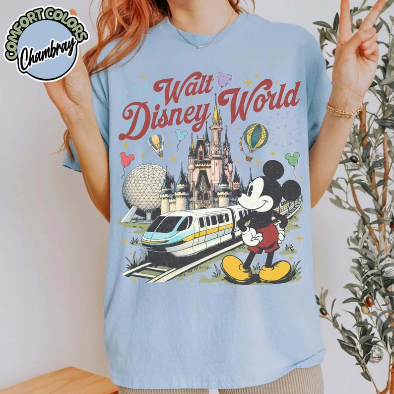 May include: Pale blue t-shirt with a Walt Disney World graphic. The design shows Cinderella's Castle, Mickey Mouse, a monorail, and hot air balloons. The text "Walt Disney World" is in a retro style.