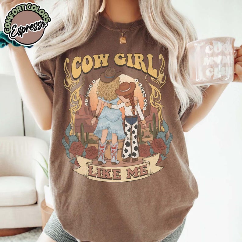 May include: A dark brown t-shirt featuring a graphic of two figures in cowboy hats standing in a desert landscape with cacti and roses. The text reads COW GIRL and LIKE ME. The shirt has a round neckline and short sleeves, with a circular logo in the corner.