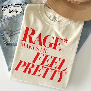 May include: An ivory short-sleeved t-shirt with a crew neckline featuring the red text RAGE* MAKES ME FEEL PRETTY printed on the front. The shirt has a Comfort Colors brand label on the inside collar and is made of cotton fabric.