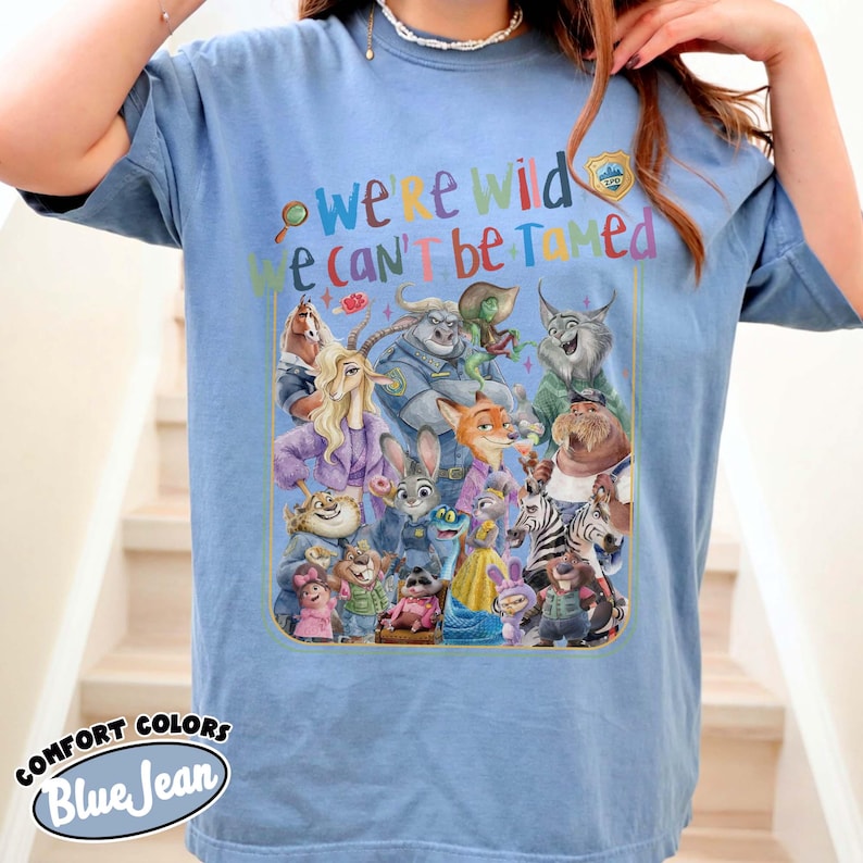 May include: Blue t-shirt with a colorful graphic featuring characters from a cartoon movie. The text "we're wild, we can't be tamed" is printed above the characters. The shirt is labeled "Comfort Colors Blue Jean".
