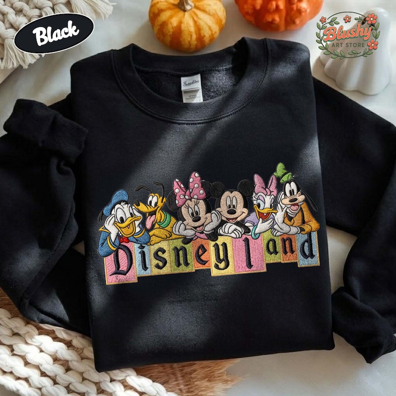May include: A black crewneck sweatshirt featuring an embroidered graphic of Disney characters including Donald Duck, Pluto, Minnie Mouse, Mickey Mouse, Daisy Duck, and Goofy. Below the characters, the word Disneyland is embroidered in colorful blocks.
