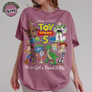 May include: A berry-colored t-shirt featuring a graphic print of Disney Pixar Toy Story characters including Woody, Buzz Lightyear, Jessie, Rex, and Forky. The design includes the text Toy Story 5 and the phrase You're Got a Friend In Me.