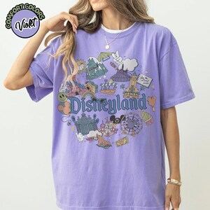 Puede incluir: Una camiseta de color violeta con un gr&aacute;fico de Disneyland. El gr&aacute;fico presenta varios iconos del parque, incluyendo un castillo, una noria y Mickey Mouse. La palabra "Disneyland" se muestra en una fuente caprichosa.