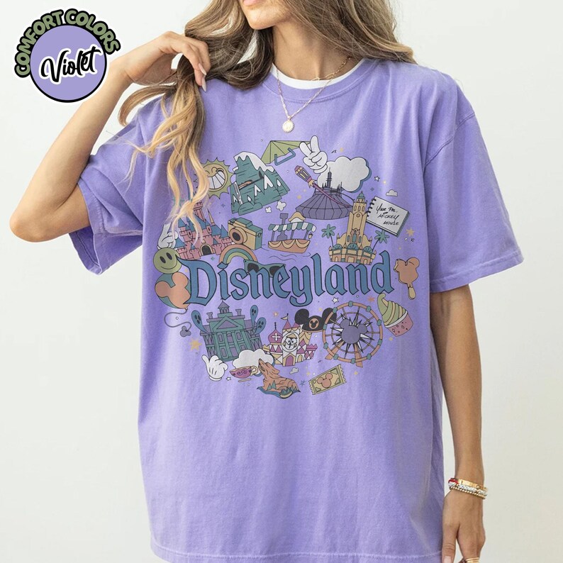 Puede incluir: Una camiseta de color violeta con un gr&aacute;fico de Disneyland. El gr&aacute;fico presenta varios iconos del parque, incluyendo un castillo, una noria y Mickey Mouse. La palabra "Disneyland" se muestra en una fuente caprichosa.