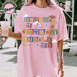 May include: A light pink t-shirt featuring a graphic of colorful alphabet blocks spelling out the text YOU ARE A COWBOY LIKE ME. The design includes illustrations of a rocket, a cowboy hat, a sheriff star, and a pair of brown cowboy boots.