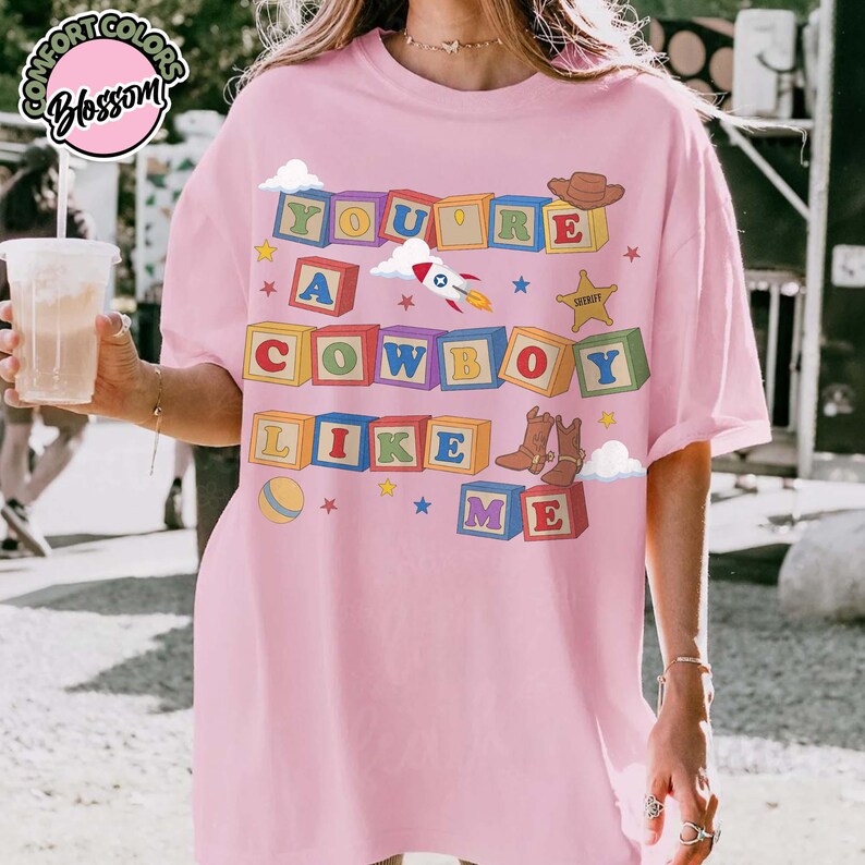 May include: A light pink t-shirt featuring a graphic of colorful alphabet blocks spelling out the text YOU ARE A COWBOY LIKE ME. The design includes illustrations of a rocket, a cowboy hat, a sheriff star, and a pair of brown cowboy boots.