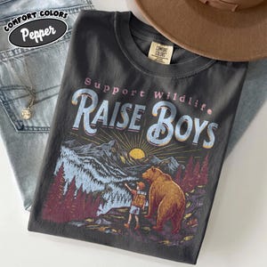 May include: A charcoal grey t-shirt featuring a graphic print of a bear and a child standing in a forest landscape with mountains and a sun. The text reads Support Wildlife RAISE BOYS. The shirt has a Comfort Colors label at the inner neckline.