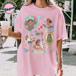 May include: A light pink t-shirt featuring a colorful graphic print of characters from the movie Moana, including Maui, Moana in a boat, Hei Hei, Pua, and Te Fiti, with the text Moana & Friends displayed at the bottom of the central design.
