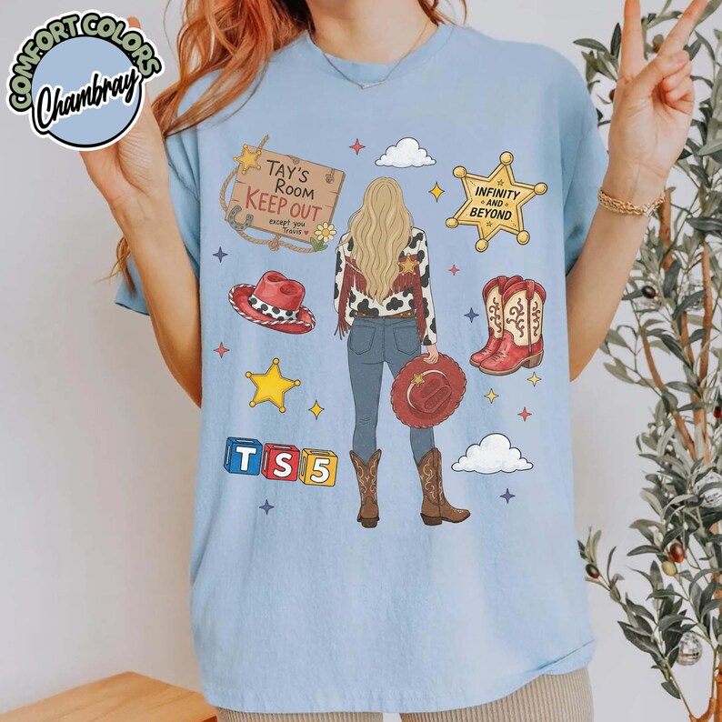 May include: A light blue t-shirt featuring a graphic of a person with long blonde hair wearing a cowgirl outfit. The design includes a sign reading Tay's Room Keep Out, a star labelled Infinity and Beyond, cowboy boots, hats, and blocks with TS5.