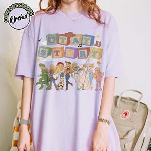 May include: A light purple short-sleeve t-shirt featuring a graphic print of colourful alphabet blocks spelling out TAY STORY above a line of illustrated characters in various outfits, including a dinosaur costume and a space-themed suit.