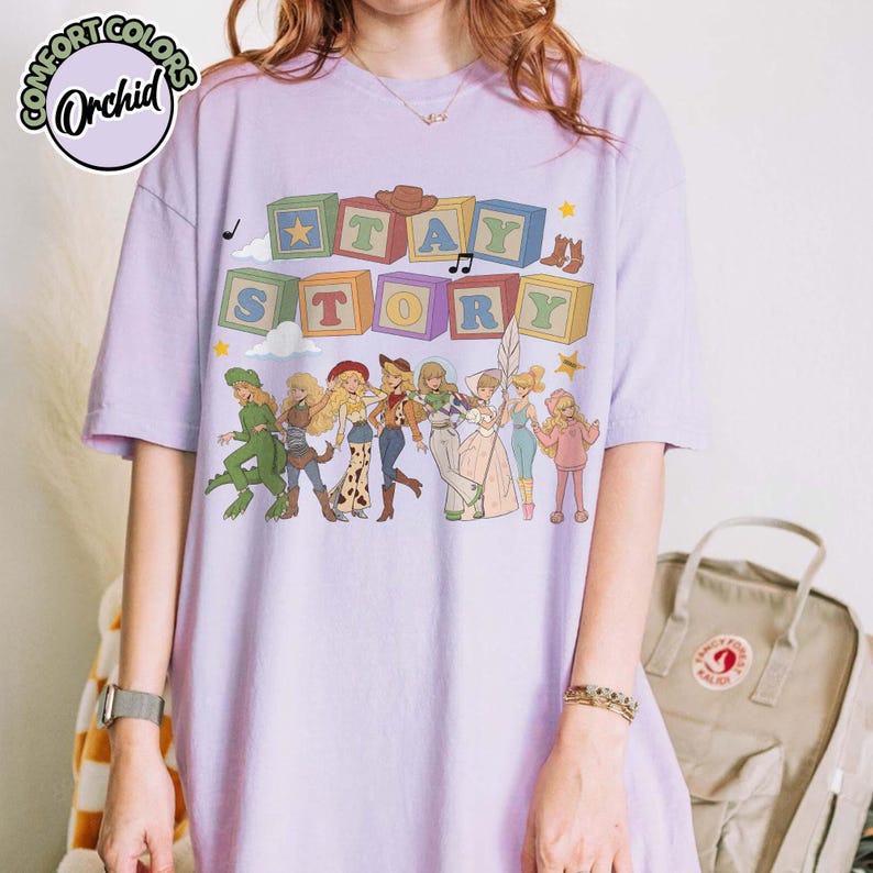 May include: A light purple short-sleeve t-shirt featuring a graphic print of colourful alphabet blocks spelling out TAY STORY above a line of illustrated characters in various outfits, including a dinosaur costume and a space-themed suit.