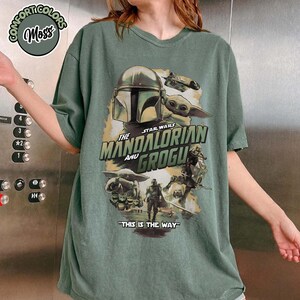 May include: A moss green short-sleeve t-shirt featuring a graphic print of The Mandalorian and Grogu. The design includes the text "The Mandalorian and Grogu" and "This is the way" along with various character illustrations from the Star Wars series.