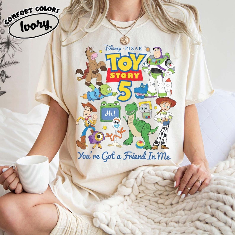 May include: An ivory t-shirt featuring a colorful graphic of Disney Pixar Toy Story 5 characters including Woody, Buzz Lightyear, Jessie, Rex, and Forky. The design includes the text Toy Story 5 and the phrase You've Got a Friend In Me.
