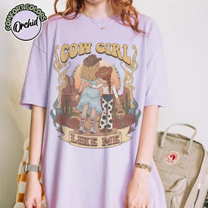 May include: A light purple t-shirt featuring a graphic of two figures in cowboy hats standing in a desert landscape with cacti and roses. The text reads COW GIRL and LIKE ME, with the phrase I KNEW YOU I KNEW YOU curving around the figures.