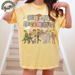 May include: A pale yellow t-shirt featuring a graphic of colourful alphabet blocks spelling out "TAY STORY" above a line of illustrated characters dressed in various costumes, including a dinosaur, a cowgirl, and a space ranger, with musical notes.
