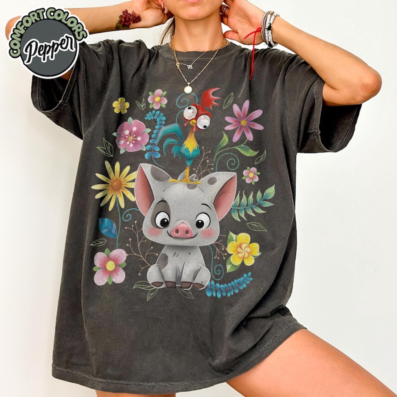 May include: Dark gray t-shirt featuring a cartoon pig with a rooster on its head, surrounded by colorful flowers and foliage. The shirt has the text "Comfort Colors Pepper" in a circle on the upper left.