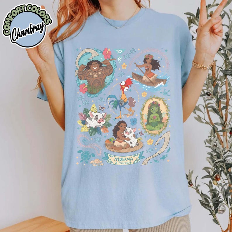 May include: A light blue t-shirt featuring a colorful graphic print of characters from the movie Moana, including Maui, Moana, Hei Hei, Pua, and Te Fiti. The design includes tropical flowers, leaves, and the text Moana & Friends.
