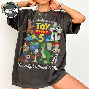 May include: A charcoal gray short-sleeve t-shirt featuring a graphic print of Disney Pixar Toy Story characters including Woody, Buzz Lightyear, Jessie, Rex, and Forky. The design includes the text Toy Story 5 and You're Got a Friend In Me.