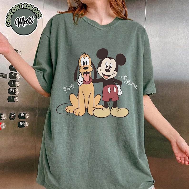 May include: Green t-shirt featuring a graphic of Mickey Mouse and Pluto. The design includes the text "Mickey Mouse" and "Pluto". The shirt has a round neckline and short sleeves. The shirt is a relaxed fit.