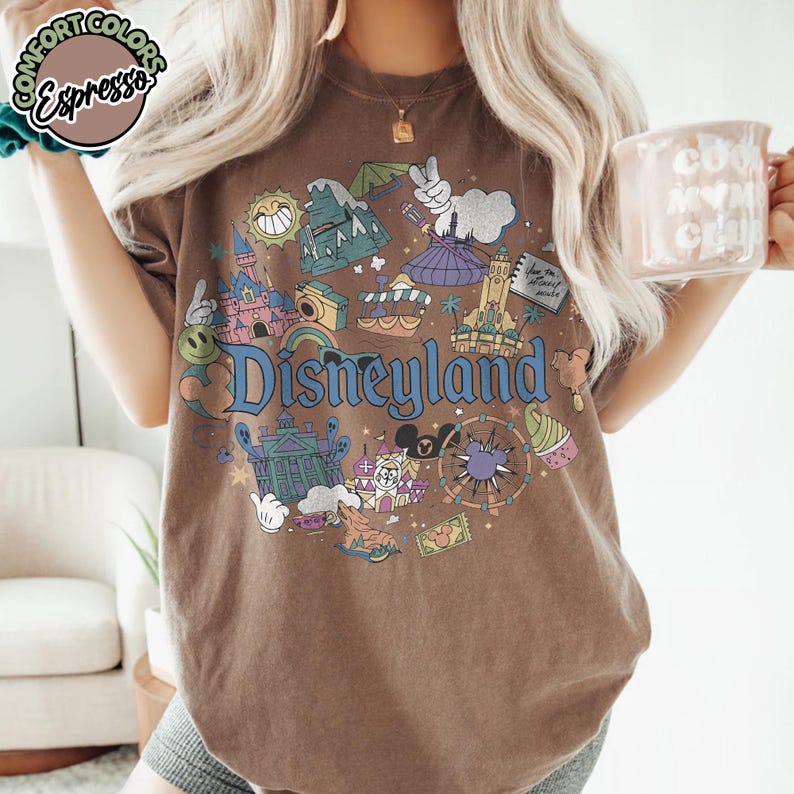 Puede incluir: Camiseta marr&oacute;n con un gr&aacute;fico de Disneyland. El gr&aacute;fico presenta varios iconos de Disneyland, incluyendo un castillo, un carrusel y una noria, en un estilo colorido y vintage. La palabra "Disneyland" se muestra prominentemente en escritura azul.