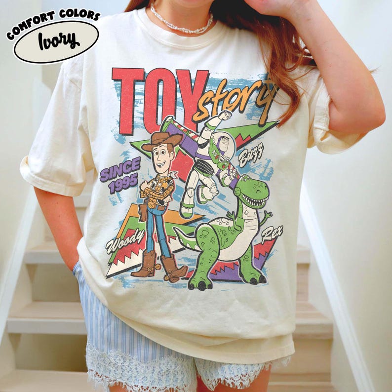 Puede incluir: Camiseta Comfort Colors color marfil con un dise&ntilde;o de Toy Story que muestra a Woody, Buzz Lightyear y Rex. La camiseta tiene el texto "TOY STORY" y "SINCE 1995". Se combina con shorts a rayas azules y blancas.