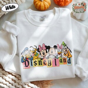 May include: A white crewneck sweatshirt featuring a colorful embroidered graphic of Disney characters including Donald Duck, Pluto, Minnie Mouse, Mickey Mouse, Daisy Duck, and Goofy. Below the characters, the word Disneyland is embroidered in black.