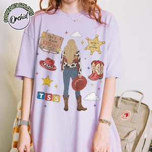 May include: A light purple t-shirt featuring a graphic print of a person with long blonde hair seen from behind, wearing a cow print shirt, blue jeans, and brown boots. The design includes a wooden sign reading Tay's Room Keep Out, a sheriff star, a red cowboy hat, red cowboy boots, and blocks spelling TS5.