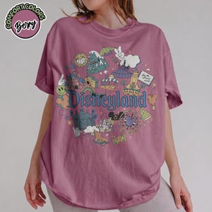 Puede incluir: Camiseta de color malva con un gr&aacute;fico de Disneyland. El gr&aacute;fico presenta varios iconos del parque, incluyendo un castillo, el Matterhorn y el logotipo de Disneyland. La camiseta tiene mangas enrolladas y un corte holgado.
