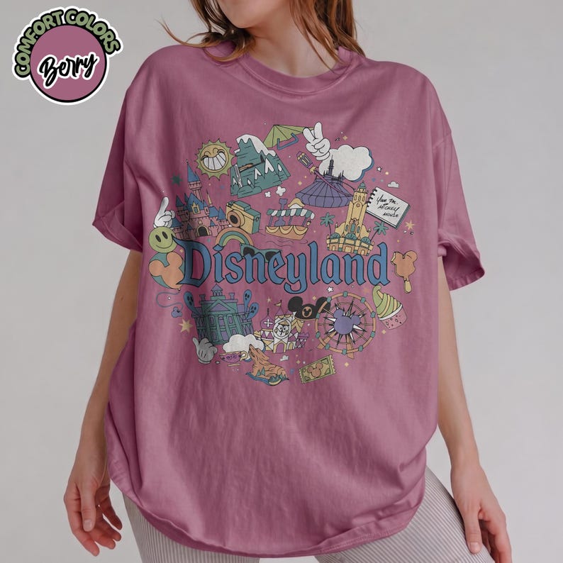 Puede incluir: Camiseta de color malva con un gr&aacute;fico de Disneyland. El gr&aacute;fico presenta varios iconos del parque, incluyendo un castillo, el Matterhorn y el logotipo de Disneyland. La camiseta tiene mangas enrolladas y un corte holgado.
