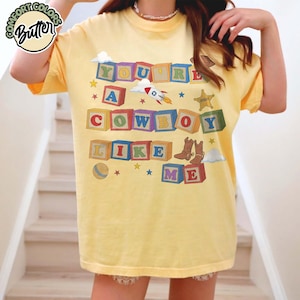 May include: A pale yellow t-shirt featuring a graphic print of colorful alphabet blocks spelling out the text YOU'RE A COWBOY LIKE ME. The design includes illustrations of a rocket, a sheriff badge, cowboy boots, a ball, clouds, and stars.