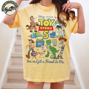 May include: A pale yellow short-sleeve t-shirt featuring a graphic print of Disney Pixar Toy Story characters including Woody, Buzz Lightyear, Jessie, Rex, and Forky. The text reads Toy Story 5 and You've Got a Friend In Me. A Comfort Colors Butter logo is visible.