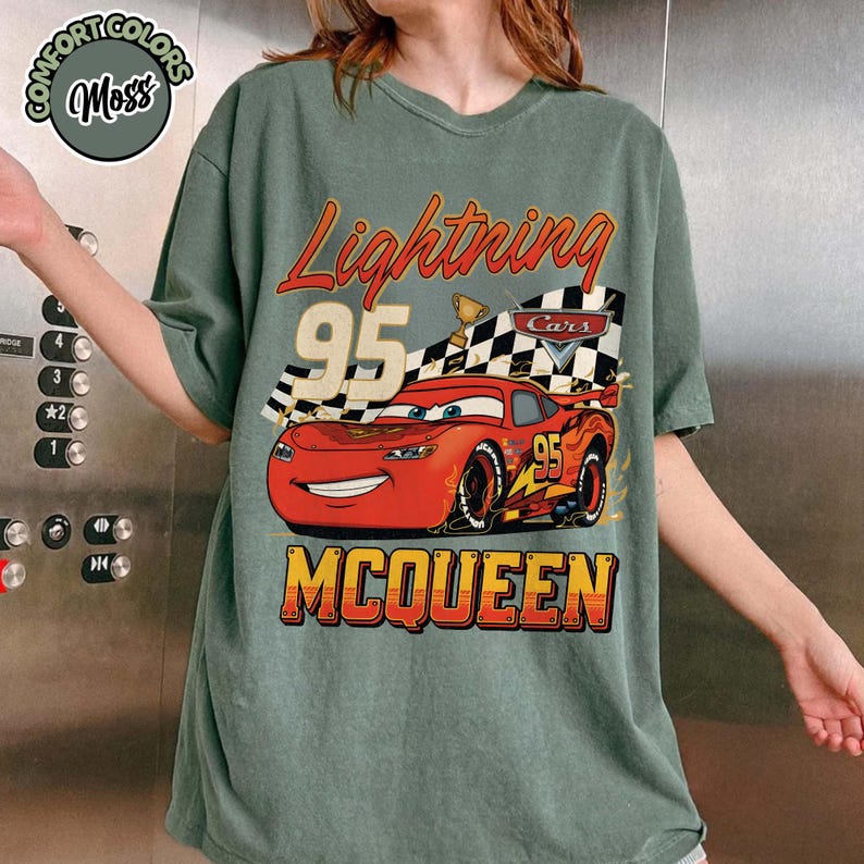 Pu&ograve; includere: T-shirt verde con grafica di Saetta McQueen del film Cars. Il design include la scritta "Lightning 95 McQueen" con una bandiera a scacchi e un trofeo. La maglietta ha un aspetto vintage e usurato.