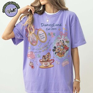 Peut inclure: Un t-shirt violet Comfort Colors avec des graphismes de Disneyland. Le design comprend Mickey Mouse, Minnie Mouse, Dingo et Donald Duck. Le t-shirt porte &eacute;galement l'inscription "Disneyland Est. 1955" et d'autres illustrations sur le th&egrave;me de Disney.