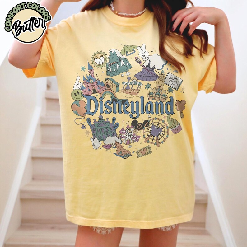 Puede incluir: Camiseta amarilla con un gr&aacute;fico de Disneyland. El dise&ntilde;o presenta varios iconos de Disneyland, incluyendo un castillo, una noria y el logotipo de Disneyland. La camiseta tiene mangas cortas y un corte relajado, perfecta para un d&iacute;a casual.
