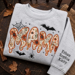 Könnte beinhalten: Ein hellgrauer Rundhals-Pullover mit dem Wort MAMA in einer tropfenden Schriftart mit Blumenmuster in Orange, Creme und Grün. Das Design umfasst schwarze Stickereien von Spinnennetzen, Fledermäusen und einer Spinne. Auf dem Ärmel sind die Namen Ethan, Sophia und Kaity eingestickt.