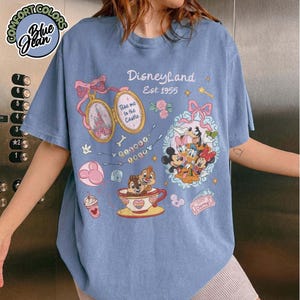 Peut inclure: T-shirt bleu clair avec un motif Disneyland. Le design comprend le texte "Disneyland Est. 1955" et des personnages Disney. Le t-shirt porte &eacute;galement l'inscription "Take me to the Castle".