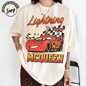 Pu&ograve; includere: T-shirt color avorio con grafica di Saetta McQueen del film Cars. Il design include la scritta "Lightning 95 McQueen", una bandiera a scacchi e un trofeo. La maglietta &egrave; del marchio Comfort Colors.