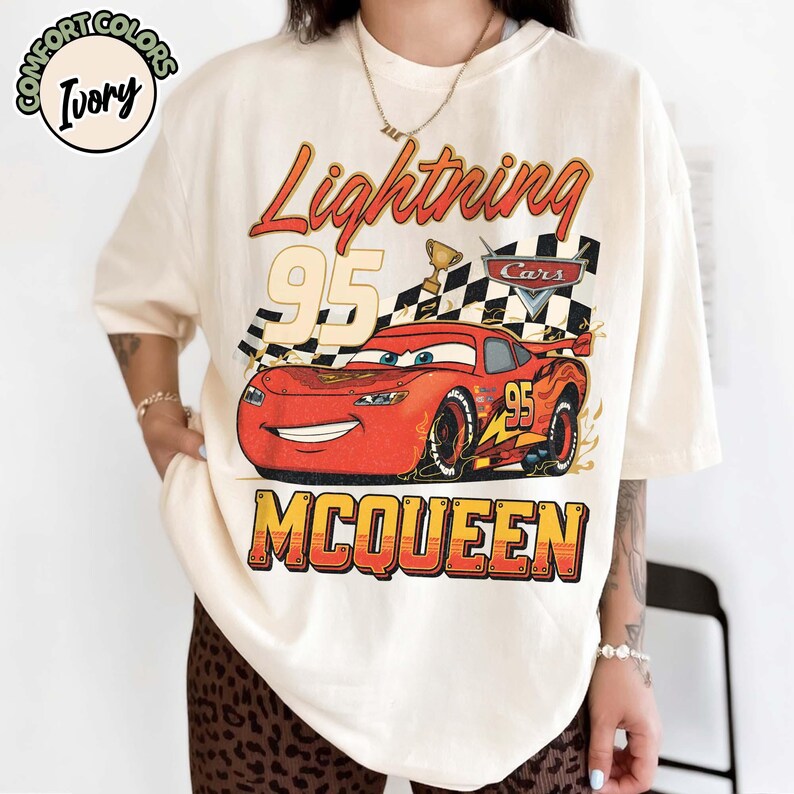 Pu&ograve; includere: T-shirt color avorio con grafica di Saetta McQueen del film Cars. Il design include la scritta "Lightning 95 McQueen", una bandiera a scacchi e un trofeo. La maglietta &egrave; del marchio Comfort Colors.